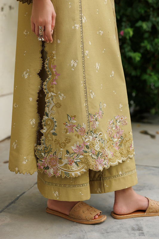 A close-up of a person wearing a mustard yellow embroidered kurta and matching wide-leg trousers, with floral embroidery concentrated along the hem and side slit. The person is wearing tan slide sandals, standing outdoors on a stone floor with greenery in the background.
